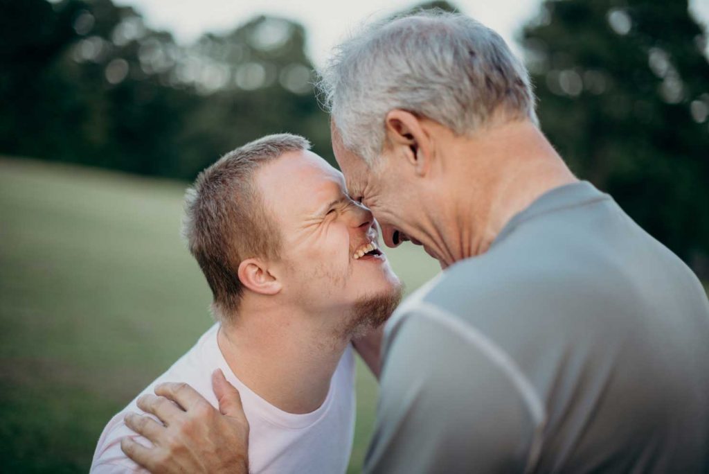 A close moment between a grey-haired father with his adolescent son who has Down Syndrom. Father and son laughing togetherm their foreheads touching.