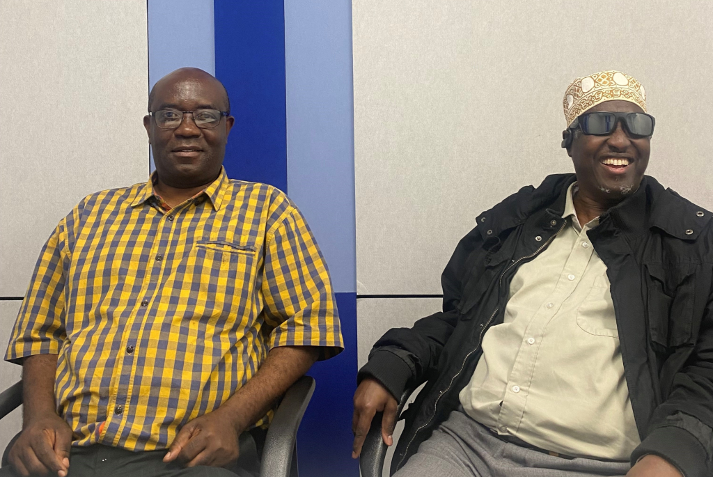 Photo of Etiene and Siyat sitting next to each other and smiling at the camera. Etiene wears glasses and a yellow checked shirt. Dr Siyat is wearing a traditional hat, sunglasses, a beige button up shirt and black jacket.