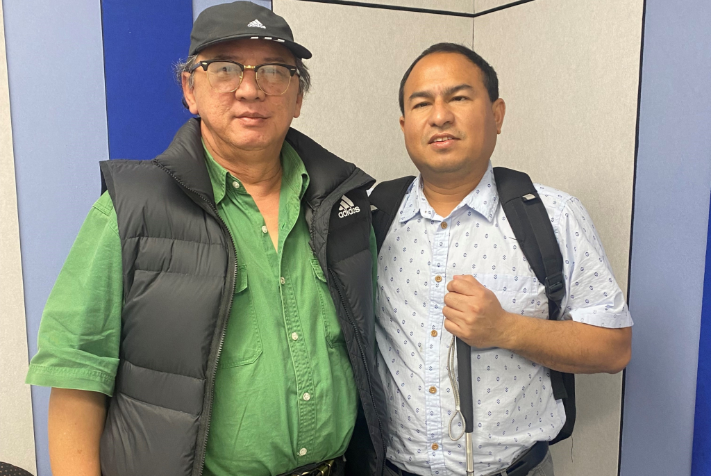 Image of two males standing together and looking at camera. One is wearing a black hat, glasses, a green button up shirt, black puffer vest and black pants. The other man is wearing a backpack, blue button up shirt, grey trousers and is holding a cane.