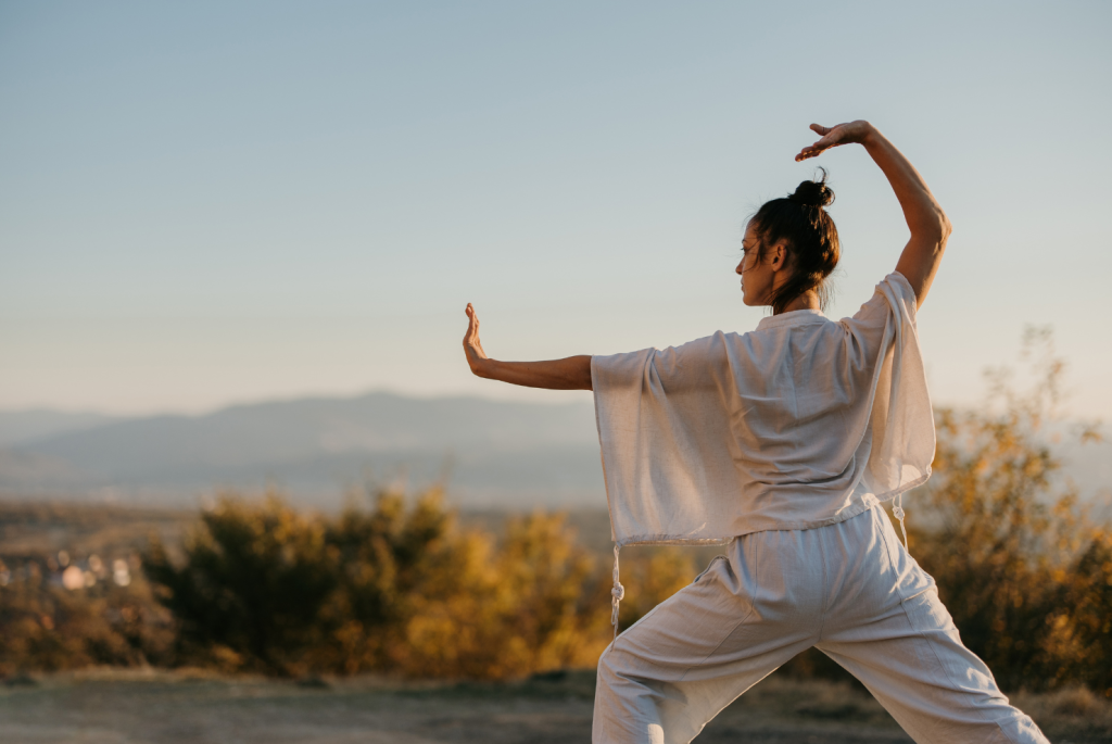 Image of a woman wearing all white who is outside and preforming a tai chi move.