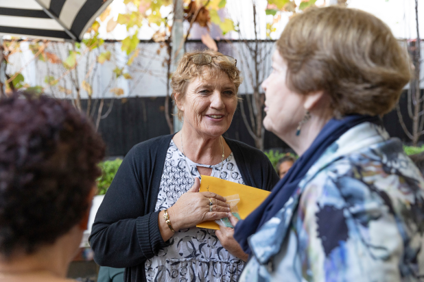 Image of a lady smiling and talking to another lady in the frame. She is wearing glasses on her head and wearing a blue and white designed top with a navy cardigan.